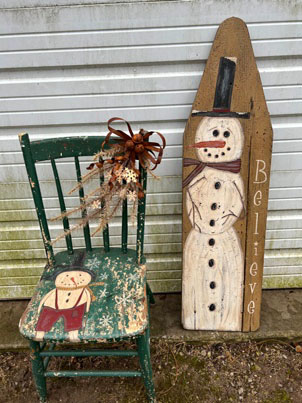 Old wooden green chair with rusty bow and a snowman in overalls painted on the seat. Old wooden ironing board with tall snowman and Believe painted on it.