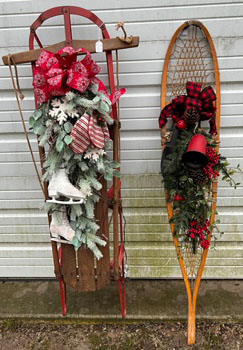 Old wooden sled with skates, greenery, and red bow on left. Snowshoe with red bell, greens, and red and black plaid ribbon at top.
