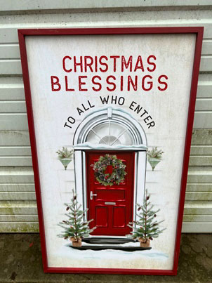 Large white sign with red trim. Painted red door with wreath and trees. Lettering says Christmas Blessings to all who enter.