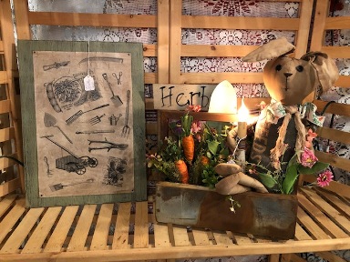 Green sign board with antique garden tools on aged paper, wood box with filled with carrots, a primitive bunny, electric candle, and flowers